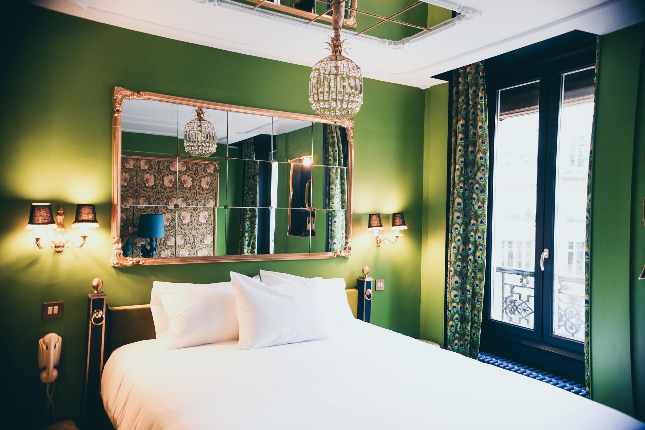 Chambre avec un lit double doré, décoration fond vert et blanc. Lampes mural, un téléphone mural, un lustre en verre et doré, un grand miroir mural, un autre sur le plafond et une grande Fênetre sécurisée par un garde-corps. 